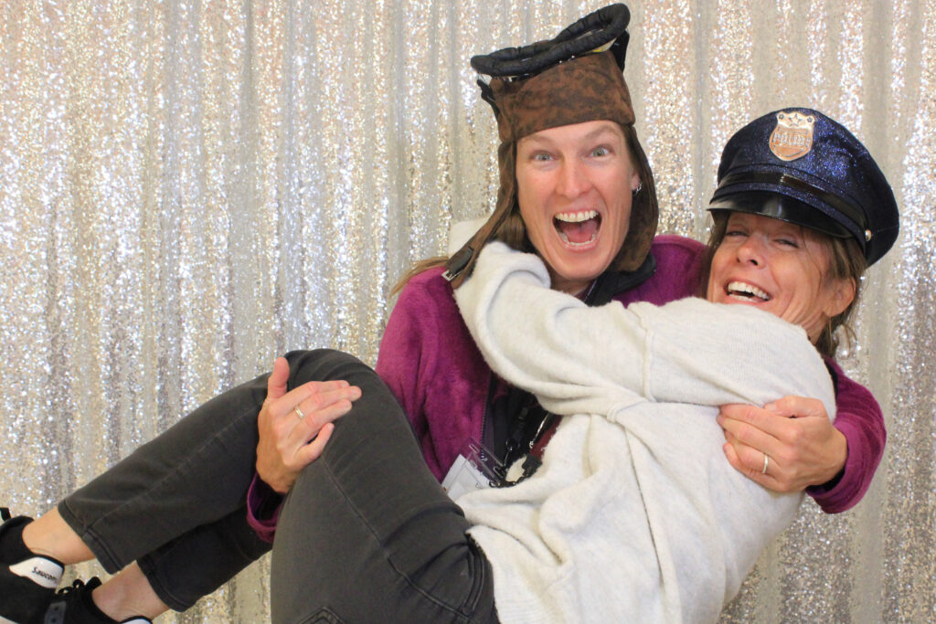 Two female Earlham faculty members pose in the photo booth wearing funny hats, one holding the other in her arms. 