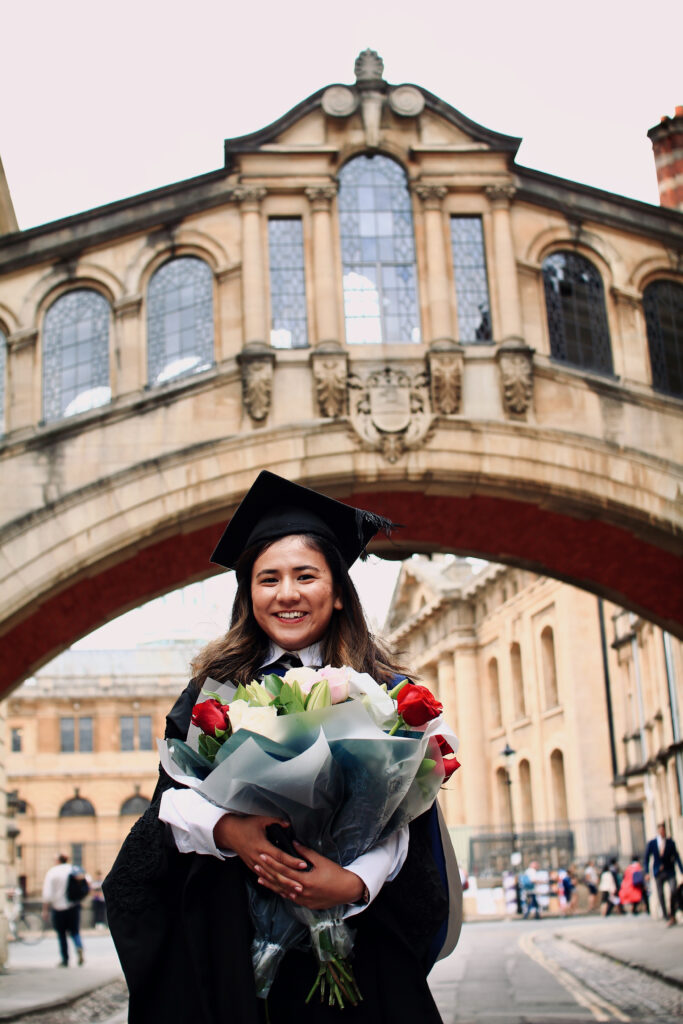 Summia Tora standing in cap and gown in London holding roses. 