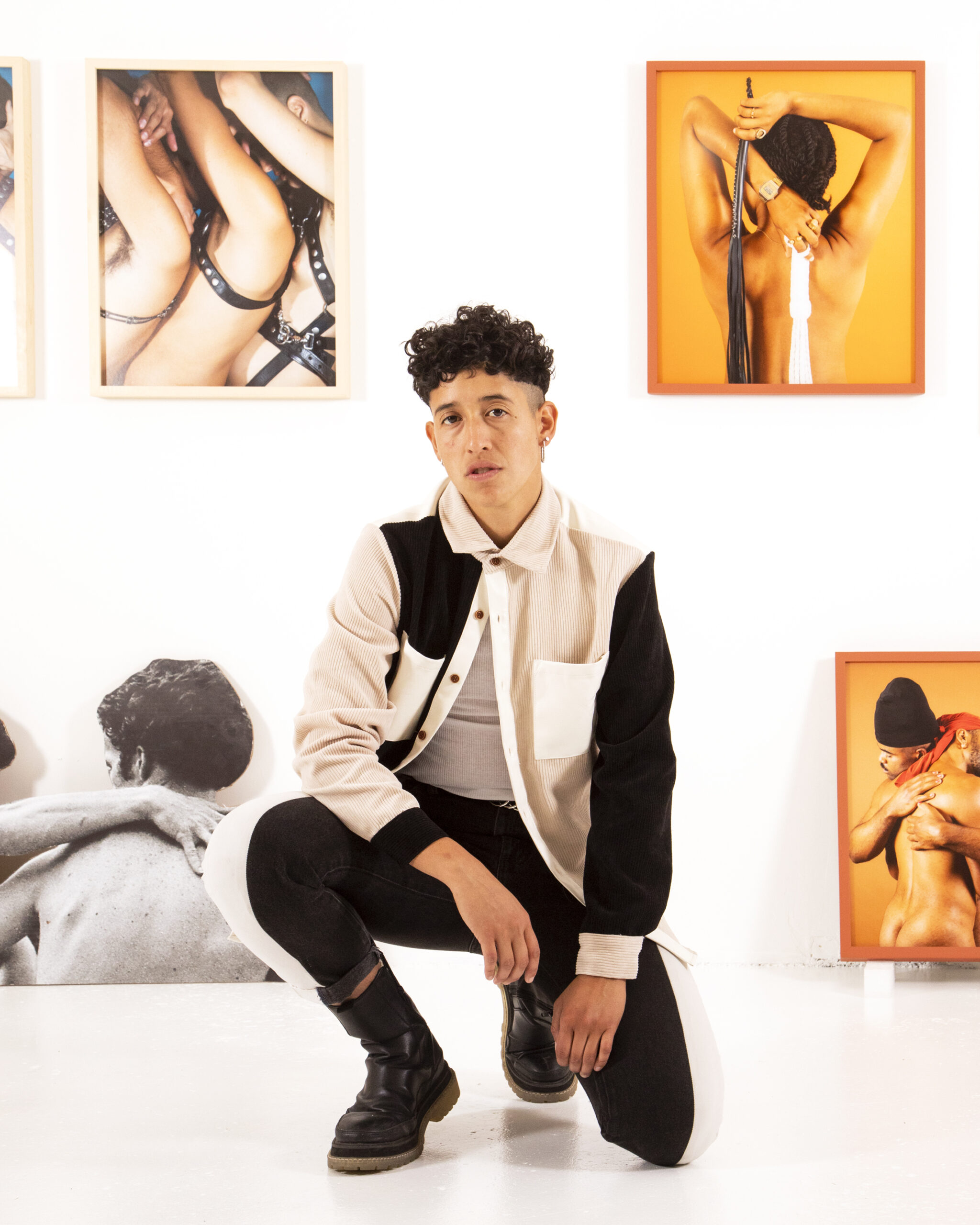 Marcel Ariza, a trans man with short curly brown hair, kneels for a pose in an art gallery