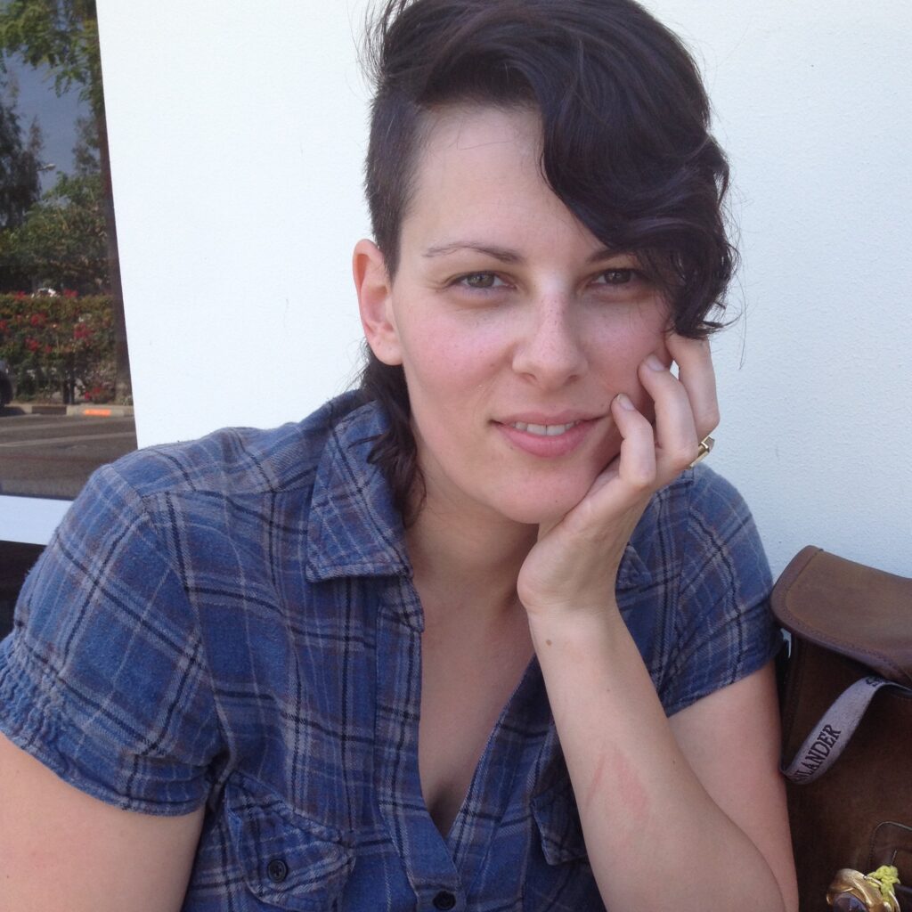 A person wearing a blue top sits staring at the camera, their hair swooped to one side, chin resting on one hand.