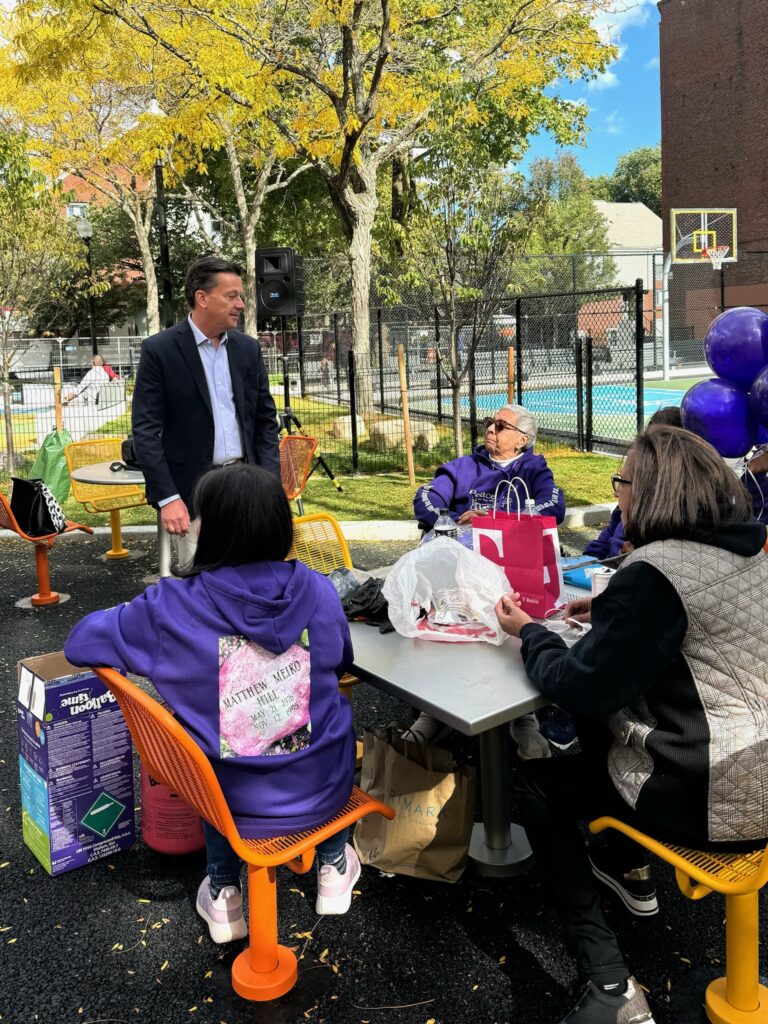 John Moran in a dark jacket and slacks addressing constituents at a public park.