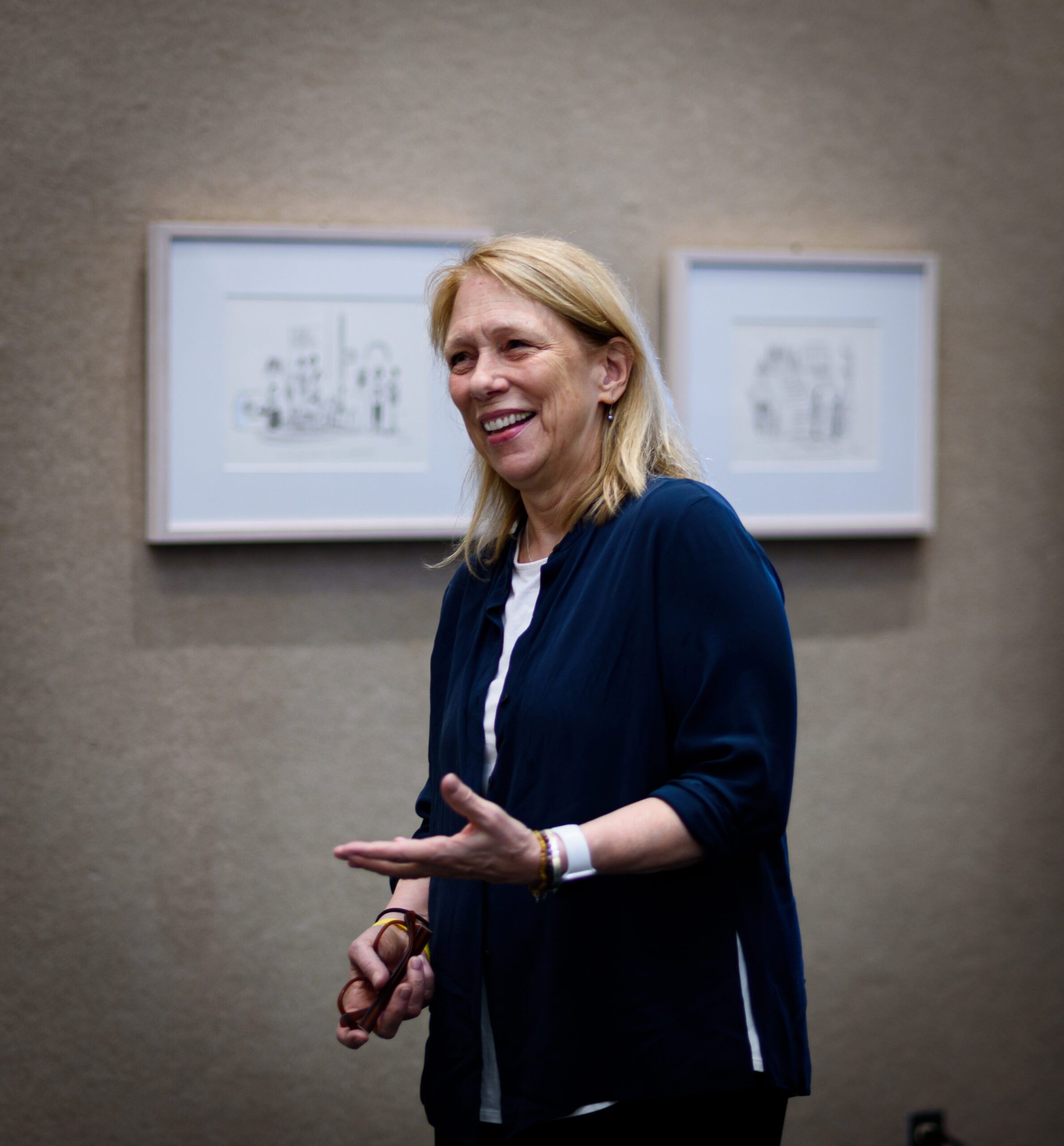 Liza Donnelly at her recent artist talk for Homecoming Weekend 2024. Liza sports blonde hair and a navy blue cardigan and stands in front of displays of her cartoons that she's drawn across the decades.