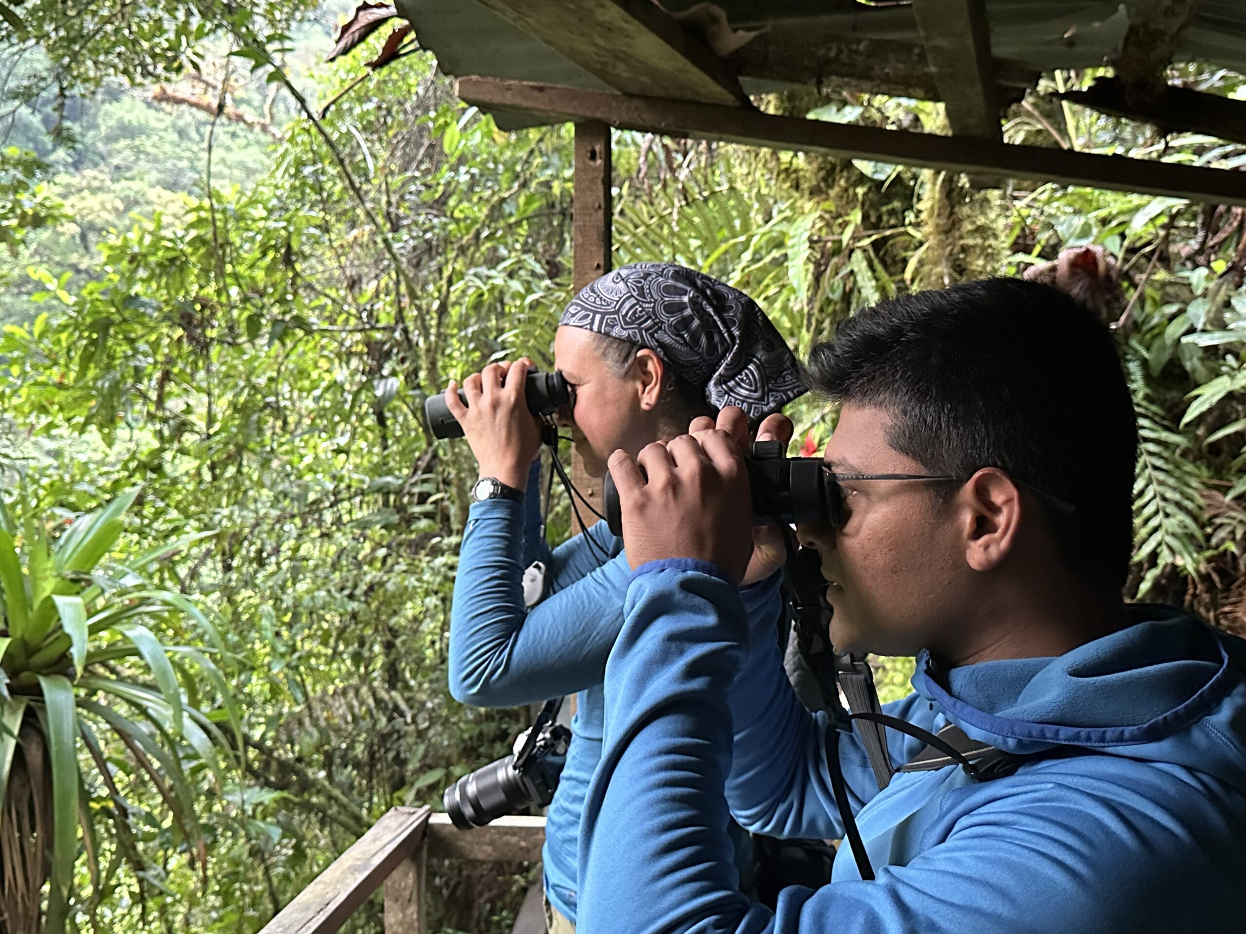 Kasun Bodawatta (right), a young adult male, watches birds through binoculars with Wendy Tori (left). Bodawatta wears a blue sweatshirt and sports wire rimmed glasses and a short black haircut. Wendy also is wearing blue and has her hair pulled back in a ponytail with a grey headband.
